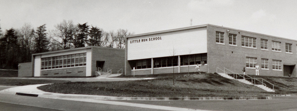 Black and white photograph of the front of Little Run Elementary School. There are a few small trees, but most of the grounds are bare grass. Two students and an adult can be seen near the building.