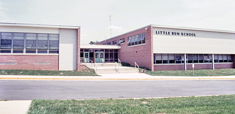 Color photograph of the front of Little Run Elementary School. The school name is visible on the front of the building. The picture appears to be from shortly after the school opened because there are no shrubs planted in front of the building that are visible in photographs from the late 1960s. A bicycle can be seen on the ground near the front steps. 